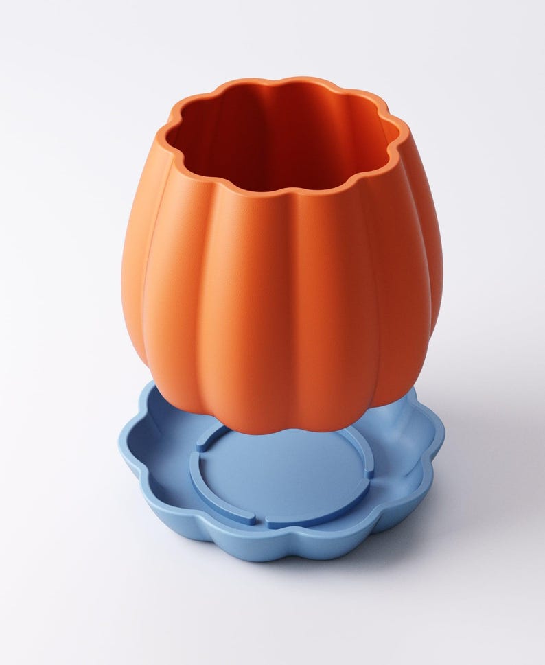 May include: An orange pumpkin-shaped container with a scalloped rim sits atop a light blue, flower-shaped base. The container has vertical ridges and a smooth, matte finish. The base has a circular indentation in the center.