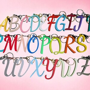 May include: A collection of colorful letter keychains, each with a different color and attached to a keyring. The letters are in a cursive font and arranged in rows against a pink background.