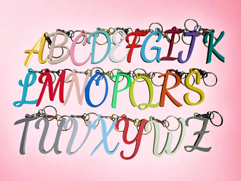 May include: A collection of colorful letter keychains, each with a different color and attached to a keyring. The letters are in a cursive font and arranged in rows against a pink background.