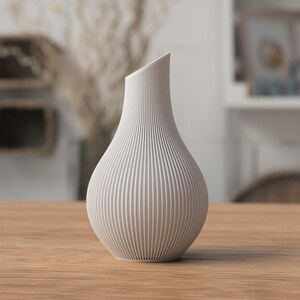 May include: A modern, off-white vase with a textured, ribbed design. The vase has a unique, angled top and a rounded base. It is displayed on a wooden table, showcasing its contemporary aesthetic. This decorative piece is suitable for home decor.