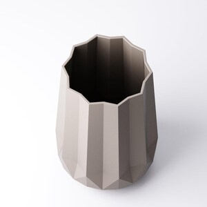 K&ouml;nnte beinhalten: Eine taupefarbene, geometrische Vase mit facettiertem Design. Die Vase hat einen breiten, gewellten Rand und einen sich verj&uuml;ngenden K&ouml;rper. Der Innenraum ist dunkel und kontrastiert mit dem hellen &Auml;u&szlig;eren. Diese dekorative Vase eignet sich f&uuml;r die Inneneinrichtung.