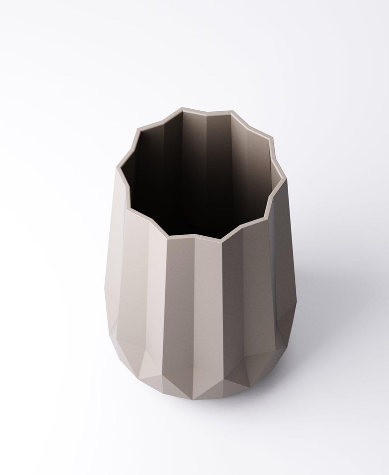 K&ouml;nnte beinhalten: Eine taupefarbene, geometrische Vase mit facettiertem Design. Die Vase hat einen breiten, gewellten Rand und einen sich verj&uuml;ngenden K&ouml;rper. Der Innenraum ist dunkel und kontrastiert mit dem hellen &Auml;u&szlig;eren. Diese dekorative Vase eignet sich f&uuml;r die Inneneinrichtung.