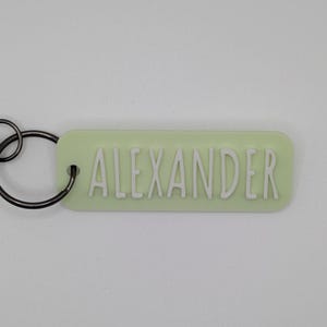 May include: A light green rectangular keychain with the name "ALEXANDER" in white, raised letters. The keychain has a black metal ring attached for keys.