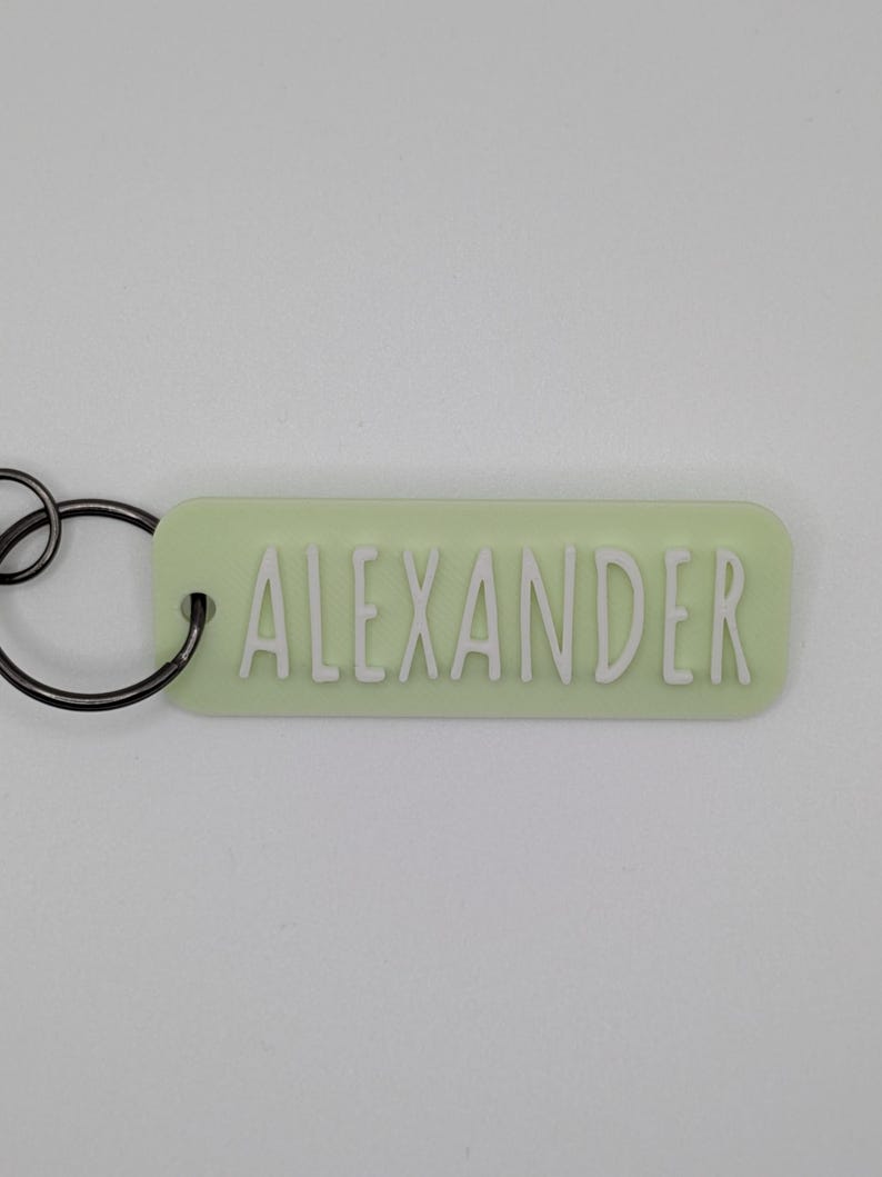 May include: A light green rectangular keychain with the name "ALEXANDER" in white, raised letters. The keychain has a black metal ring attached for keys.