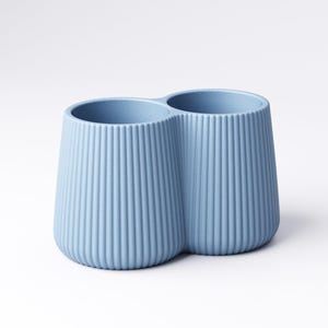 May include: A light blue, double-cup holder with vertical ribbed detailing. The two cups are connected, creating a unique shape. The holder is made of a solid material and sits on a white surface.