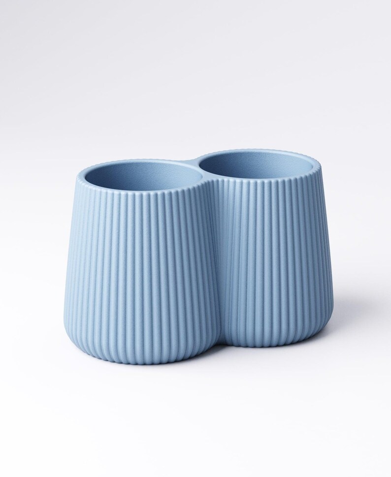 May include: A light blue, double-cup holder with vertical ribbed detailing. The two cups are connected, creating a unique shape. The holder is made of a solid material and sits on a white surface.