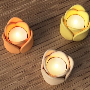 May include: Three lit, flower-shaped candle holders in shades of orange, yellow, and cream. Each holder has a small, round candle inside, casting a warm glow. The holders are arranged on a wooden surface.