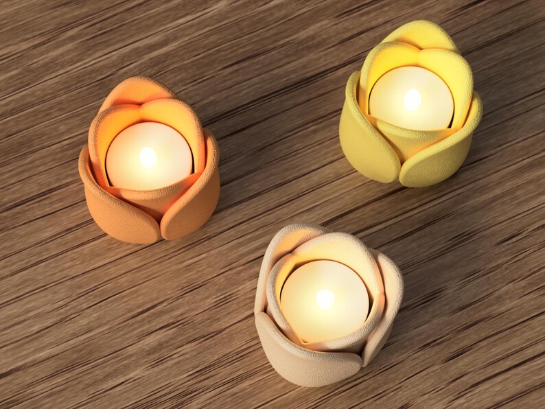 May include: Three lit, flower-shaped candle holders in shades of orange, yellow, and cream. Each holder has a small, round candle inside, casting a warm glow. The holders are arranged on a wooden surface.