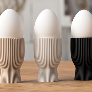 Pu&ograve; includere: Tre portauova in colori neutri, ciascuno con un uovo bianco. I portauova sono beige, grigio chiaro e nero, con un design a coste verticali. I portauova poggiano su una superficie di legno, creando un'estetica minimalista.