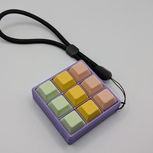 May include: A pastel-colored, nine-key keyboard with a black lanyard. The keys are arranged in a 3x3 grid, with colors including yellow, pink, and green. The keyboard is housed in a purple frame.