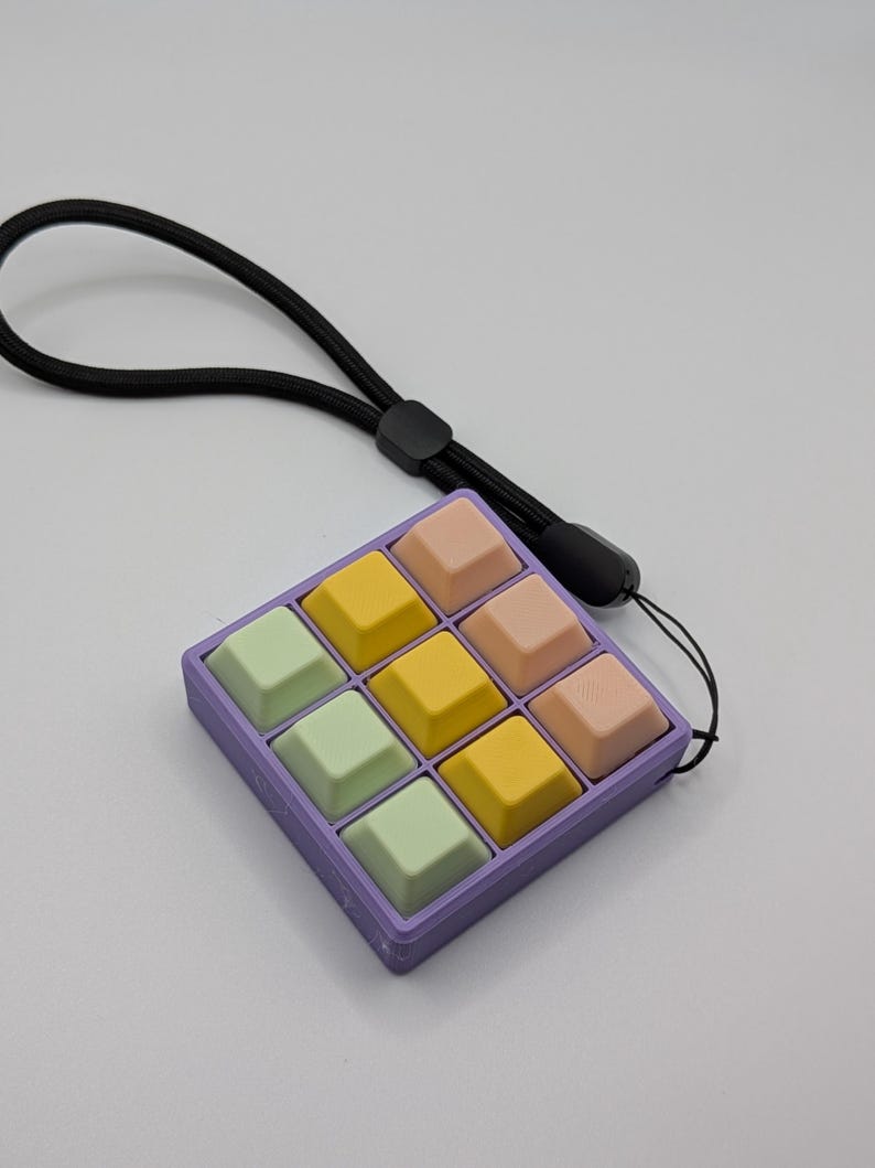 May include: A pastel-colored, nine-key keyboard with a black lanyard. The keys are arranged in a 3x3 grid, with colors including yellow, pink, and green. The keyboard is housed in a purple frame.