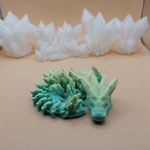 K&ouml;nnte beinhalten: Das Bild zeigt 3D-gedruckte Objekte. Mehrere wei&szlig;e, kristalline Strukturen sind im Hintergrund angeordnet. Im Vordergrund ist eine gr&uuml;n-blaue Drachenfigur zu sehen, die ein detailliertes, strukturiertes Design aufweist. Die Objekte sind vor einem neutralen Hintergrund platziert.