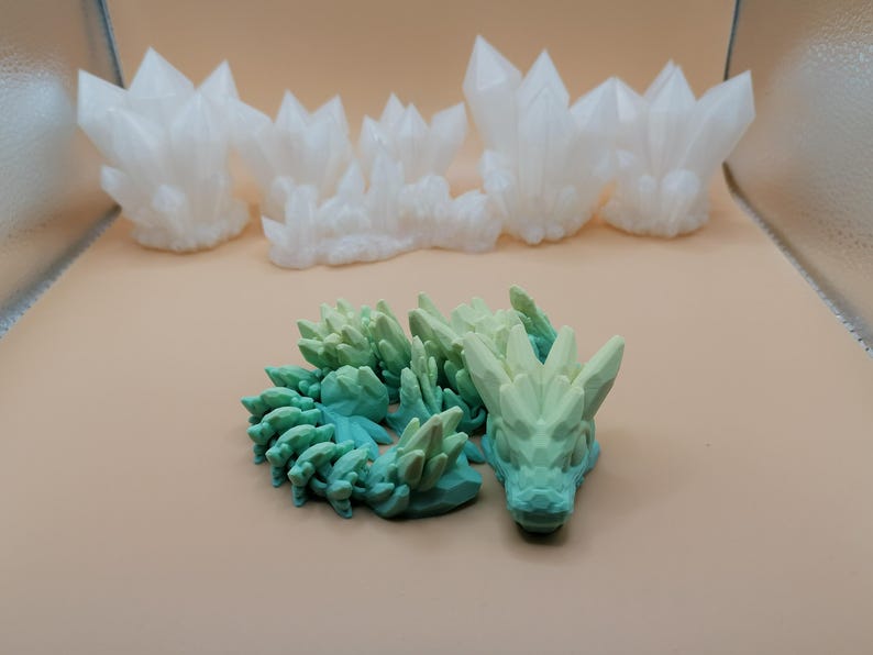 K&ouml;nnte beinhalten: Das Bild zeigt 3D-gedruckte Objekte. Mehrere wei&szlig;e, kristalline Strukturen sind im Hintergrund angeordnet. Im Vordergrund ist eine gr&uuml;n-blaue Drachenfigur zu sehen, die ein detailliertes, strukturiertes Design aufweist. Die Objekte sind vor einem neutralen Hintergrund platziert.