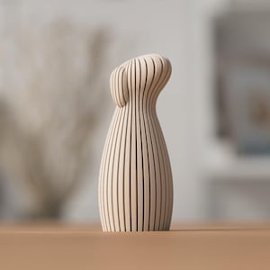 Puede incluir: Un jarr&oacute;n de madera de color claro con un dise&ntilde;o curvo &uacute;nico. El jarr&oacute;n est&aacute; construido con listones de madera verticales, creando una superficie texturizada. El jarr&oacute;n est&aacute; sobre una superficie marr&oacute;n claro, con un fondo borroso.