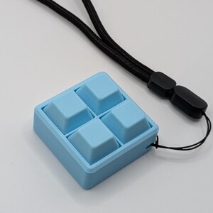 May include: A light blue square-shaped device with four raised square buttons. A black lanyard is attached to the device. The device is on a white surface.