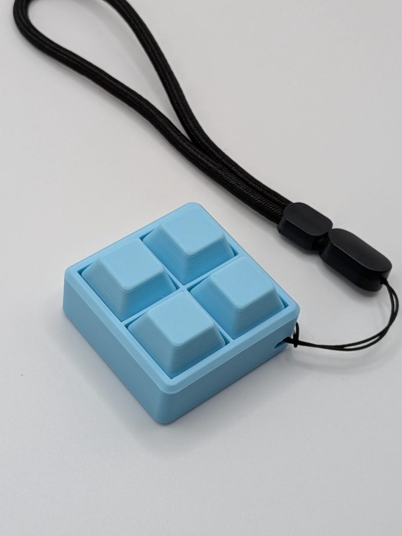 May include: A light blue square-shaped device with four raised square buttons. A black lanyard is attached to the device. The device is on a white surface.