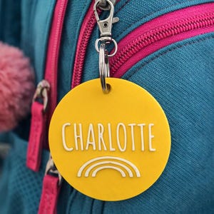 May include: Yellow circular tag with the name "CHARLOTTE" in white, and a white rainbow graphic. The tag is attached to a backpack with a silver clasp. The backpack is teal with pink zippers.