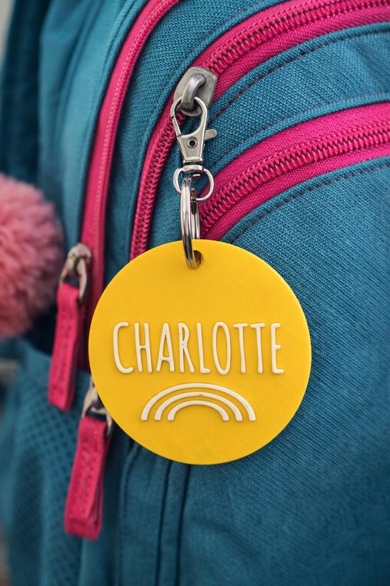 May include: Yellow circular tag with the name "CHARLOTTE" in white, and a white rainbow graphic. The tag is attached to a backpack with a silver clasp. The backpack is teal with pink zippers.