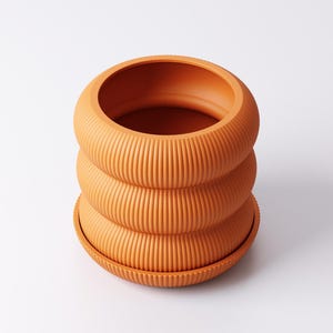 May include: A terracotta-coloured, ribbed plant pot with a matching saucer. The pot features a distinctive design with three stacked, rounded sections. The textured surface and warm colour create a modern aesthetic, suitable for indoor plants.
