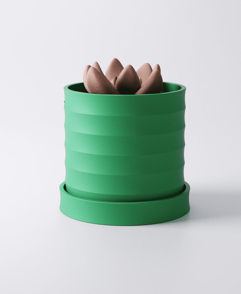 Op de afbeelding: Een groene, cilindrische plantenpot met horizontale ribbels, met een vetplant. De pot heeft een bijpassende schotel. De vetplant heeft meerdere bruine, bloembladachtige bladeren. Het ontwerp is modern en minimalistisch.