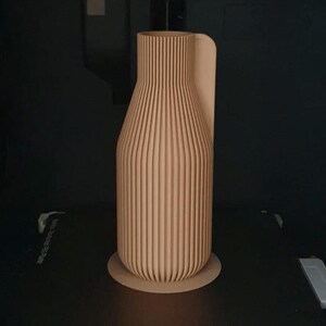 May include: A light brown, vertically ribbed vase with a rounded base and a curved handle. The vase is made of wood and sits on a circular base. The background is dark.