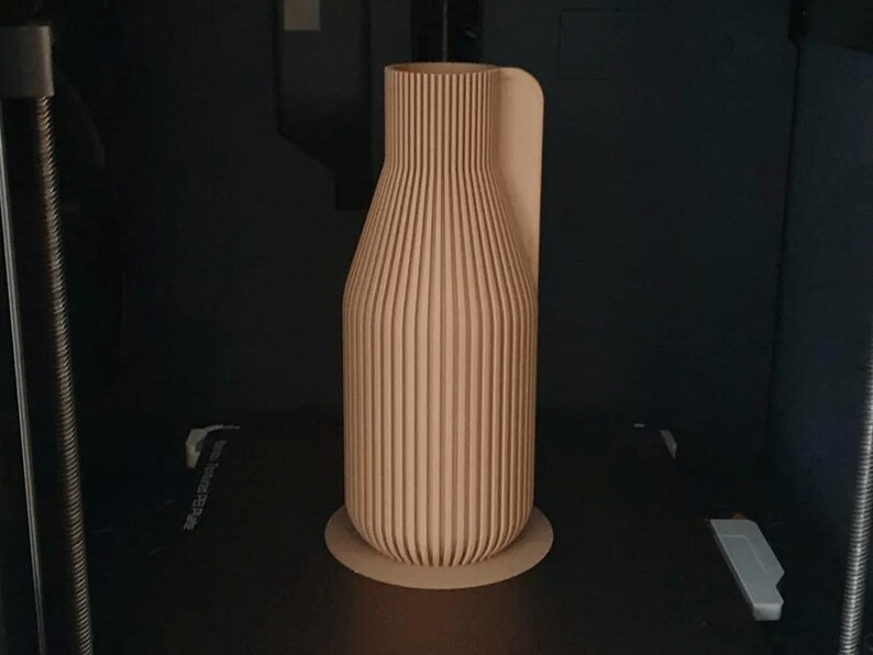 May include: A light brown, vertically ribbed vase with a rounded base and a curved handle. The vase is made of wood and sits on a circular base. The background is dark.