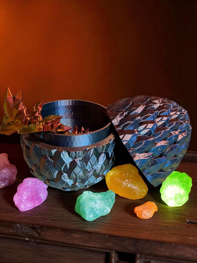 Op de afbeelding: Een blauwe decoratieve eivormige container met een getextureerd oppervlak, open om een donker interieur te onthullen. Kleurrijke, gloeiende, edelsteenachtige stenen in roze, groen, geel en oranje zijn verspreid rond de container op een houten oppervlak.