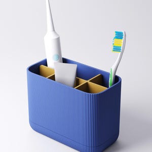 May include: A rectangular blue toothbrush holder with a ribbed exterior and gold-colored interior dividers. The holder contains a white electric toothbrush, a small white tube, and a manual toothbrush with blue, yellow, and green bristles.