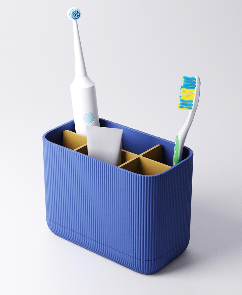 May include: A rectangular blue toothbrush holder with a ribbed exterior and gold-colored interior dividers. The holder contains a white electric toothbrush, a small white tube, and a manual toothbrush with blue, yellow, and green bristles.