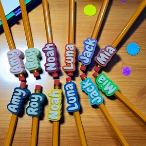 May include: A collection of yellow pencils with colorful name erasers. The erasers feature names like Amy, Roy, Noah, Luna, Jack, and Mia in various colors. The pencils are arranged on a wooden surface with colorful confetti.