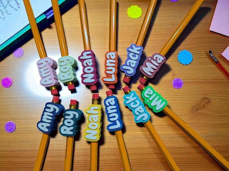 May include: A collection of yellow pencils with colorful name erasers. The erasers feature names like Amy, Roy, Noah, Luna, Jack, and Mia in various colors. The pencils are arranged on a wooden surface with colorful confetti.