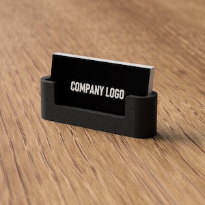 May include: A black business card holder with a black business card that reads "COMPANY LOGO" in white lettering. The holder is on a wooden surface.