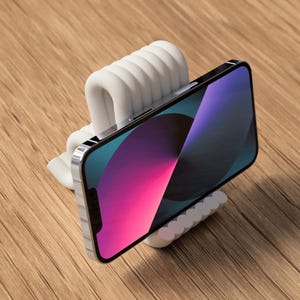 Puede incluir: Un soporte blanco y acanalado para tel&eacute;fono que sostiene un smartphone con una pantalla colorida. El soporte est&aacute; hecho de un material curvo y texturizado. El tel&eacute;fono est&aacute; inclinado, mostrando su pantalla sobre una superficie de madera.