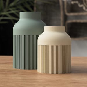 May include: Two modern ceramic vases. One is a muted green, the other is a cream color. Both have a cylindrical base with vertical ribbed detailing. The vases sit on a wooden surface.
