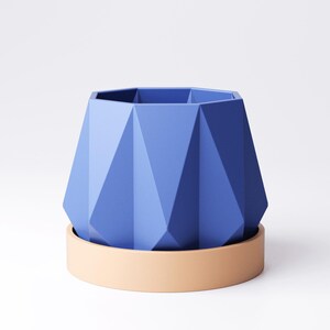 May include: A geometric, blue planter with a matching beige saucer. The planter has a faceted design and a hexagonal top. The saucer is a simple, rounded shape. The planter and saucer are made of a solid material.