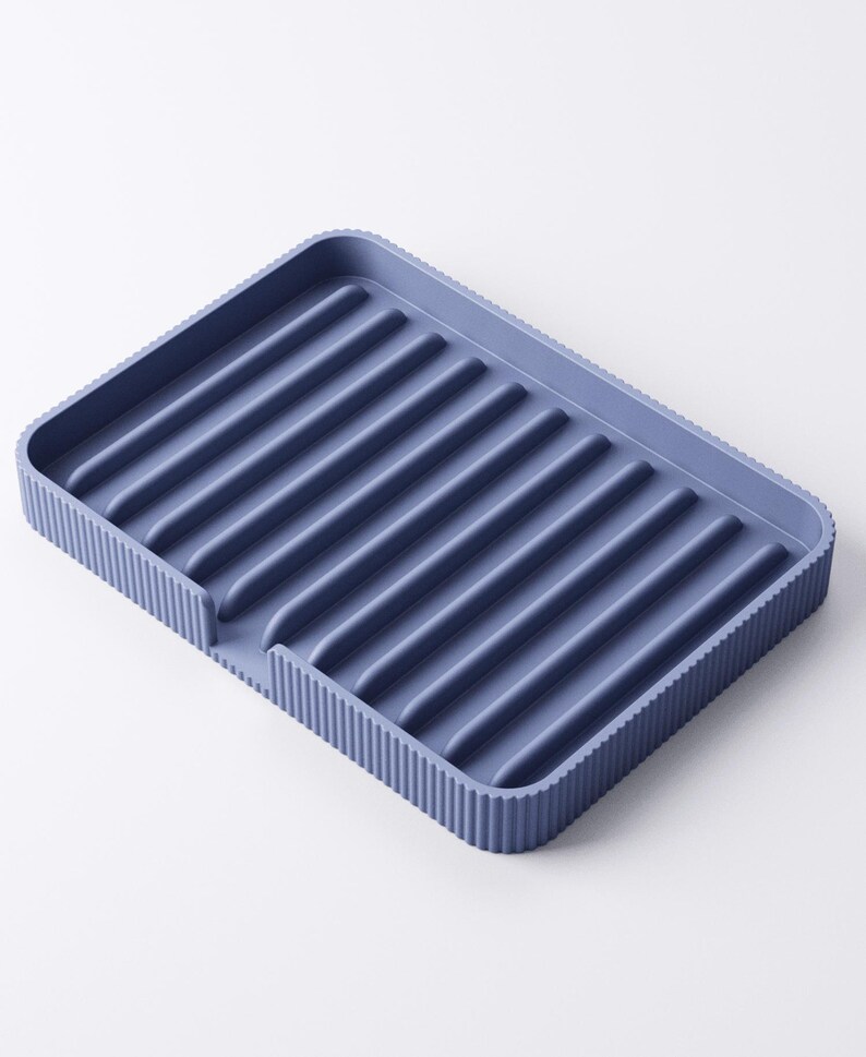 May include: A blue rectangular dish drying rack with a ribbed interior and a textured, ridged border. The rack has a small cutout on one side. The color is a muted, dusty blue.