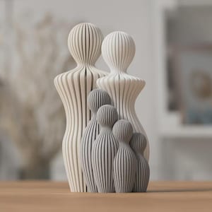 Puede incluir: Un conjunto de figuras familiares. El conjunto incluye dos figuras grandes de color crema y tres figuras m&aacute;s peque&ntilde;as de color gris. Cada figura tiene un dise&ntilde;o &uacute;nico con rayas verticales. Las figuras se exhiben sobre una superficie de madera.