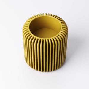 May include: A cylindrical, mustard-yellow decorative container with vertical, rounded ridges. The top edge has a series of small, vertical ridges. The interior is a matching mustard yellow. The container is set against a white background.