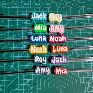May include: Clear ballpoint pens with black ink, each adorned with a colorful name tag. Names include Jack, Roy, Mia, Amy, Luna, Noah. The pens are arranged on a green cutting mat.