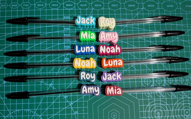 May include: Clear ballpoint pens with black ink, each adorned with a colorful name tag. Names include Jack, Roy, Mia, Amy, Luna, Noah. The pens are arranged on a green cutting mat.