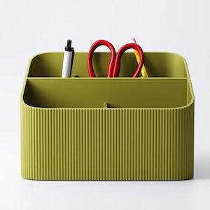 May include: Olive green desk organizer with vertical ribbed texture. The organizer holds a black pen, red scissors, and a yellow pencil. The container has two compartments and a curved top.