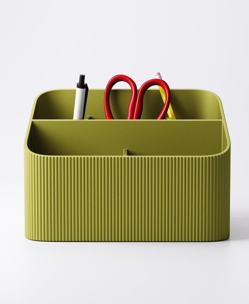 May include: Olive green desk organizer with vertical ribbed texture. The organizer holds a black pen, red scissors, and a yellow pencil. The container has two compartments and a curved top.