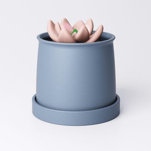 May include: A blue ceramic planter with a matching saucer holds a faux succulent. The succulent features soft pink petals with green accents. The planter has a simple, modern design, suitable for home decor.