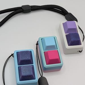 May include: Three keycap pendants in blue, pink, and purple, each with a textured bottom keycap and a smooth top keycap. The pendants are attached to black lanyards. The keycaps are housed in rectangular frames.