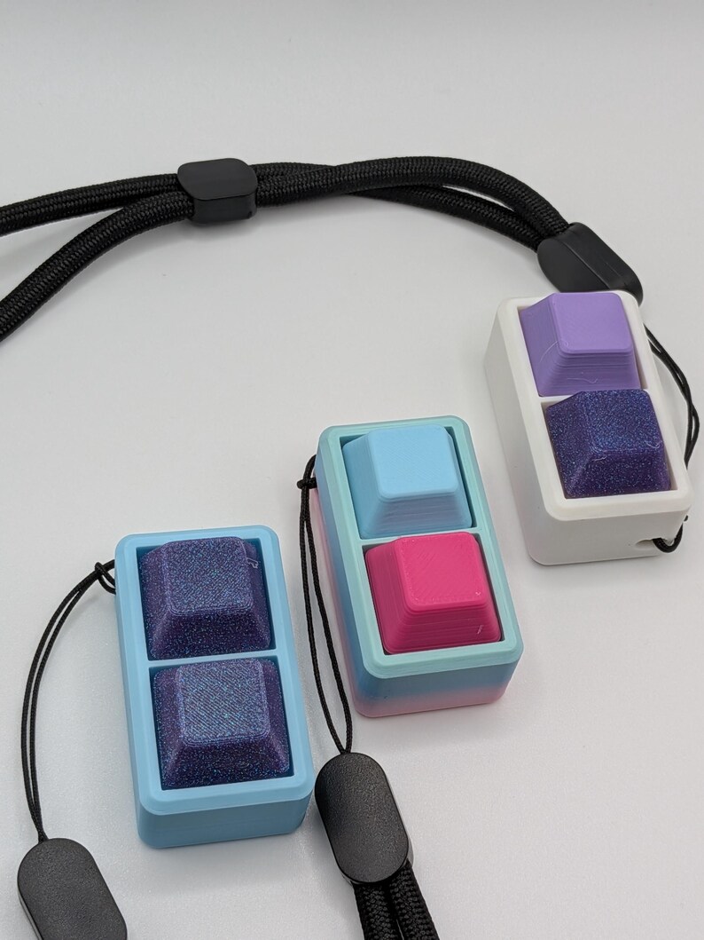 May include: Three keycap pendants in blue, pink, and purple, each with a textured bottom keycap and a smooth top keycap. The pendants are attached to black lanyards. The keycaps are housed in rectangular frames.