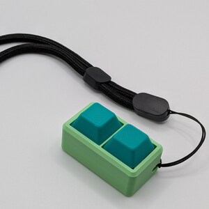 May include: A green and teal keycap set with a black lanyard. The keycaps are teal and square, set in a green rectangular frame. The black lanyard is attached to the frame.