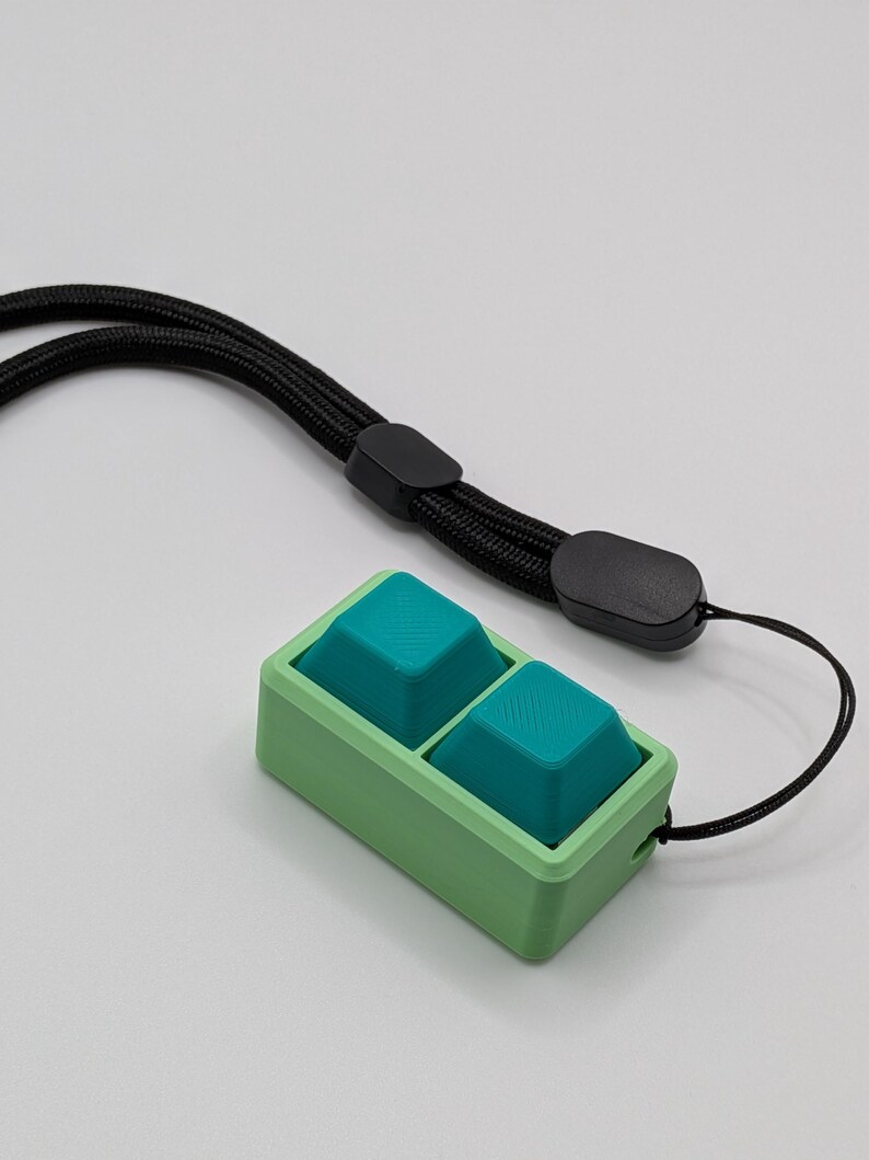 May include: A green and teal keycap set with a black lanyard. The keycaps are teal and square, set in a green rectangular frame. The black lanyard is attached to the frame.