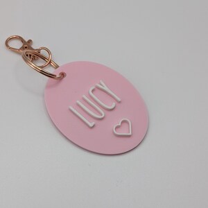May include: A pink oval keychain with the name "LUCY" in white block letters and a small white heart. The keychain has a gold-colored clasp and ring.
