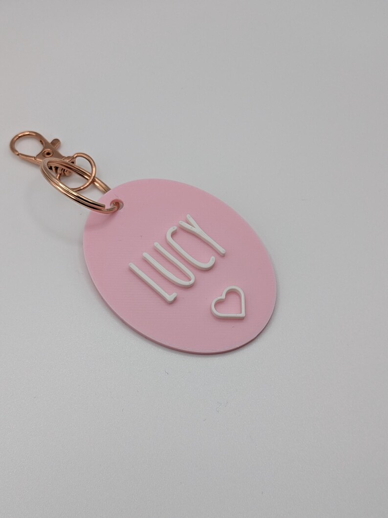 May include: A pink oval keychain with the name "LUCY" in white block letters and a small white heart. The keychain has a gold-colored clasp and ring.
