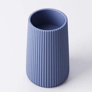 May include: A blue-gray, ribbed vase with a cylindrical shape. The vase has a slightly flared base and a textured surface with vertical lines. The top rim has a decorative, ridged edge. The vase is empty and sits on a white background.